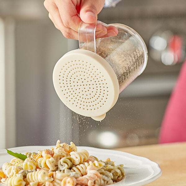 A person pouring finely ground seasoning from a Choice polycarbonate shaker onto a plate of pasta.