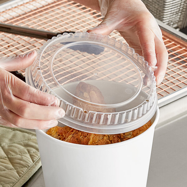 A hand in plastic gloves holding a clear plastic lid over a container of food.