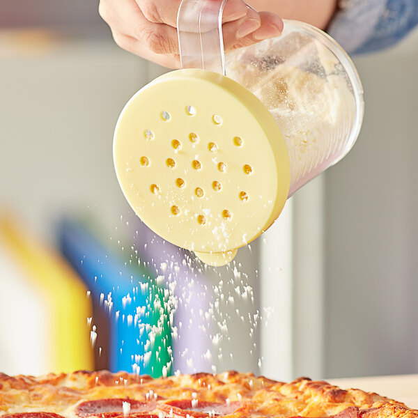 A hand pouring pepper from a yellow-lidded shaker onto a pizza.