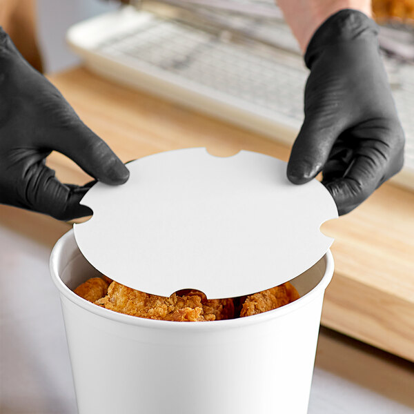 A person wearing black gloves opening a white Innopak paper bucket lid on a container of food.