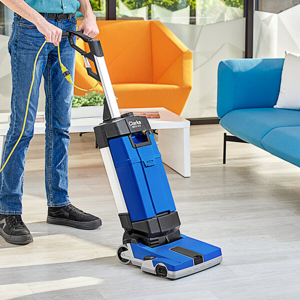A person using a Clarke walk behind cylindrical floor scrubber in a room with blue and white walls.