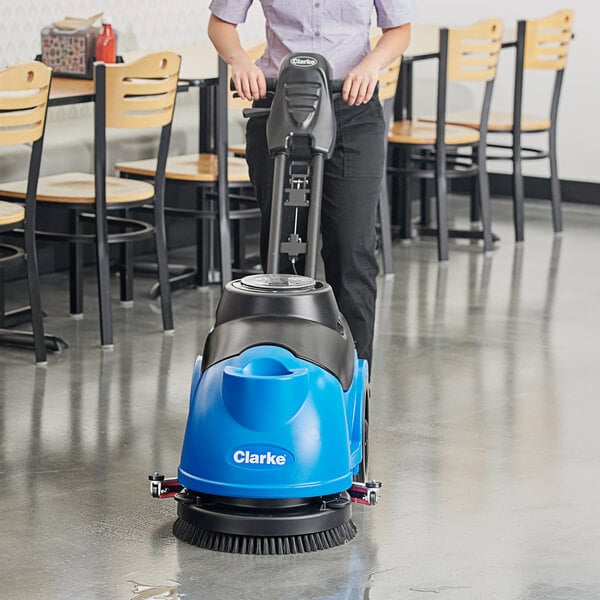 A woman using a Clarke AGM cordless walk behind floor scrubber to clean a floor in a room with tables and chairs.