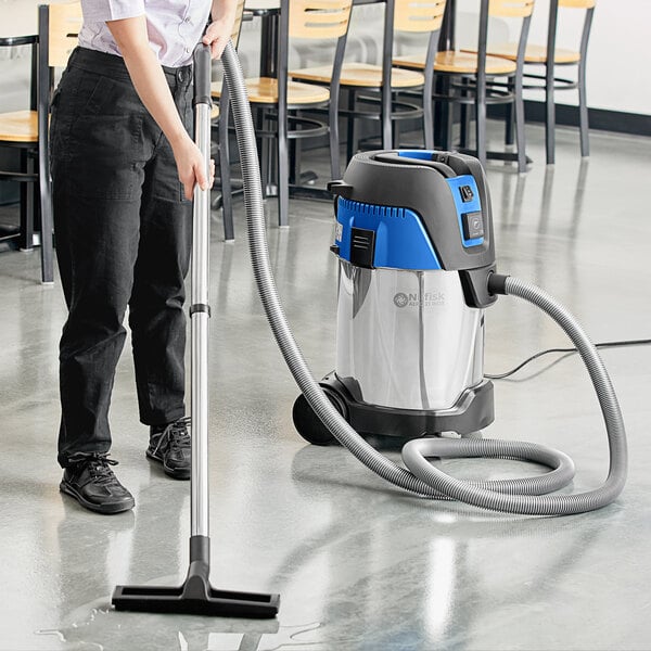 A person using a Nilfisk stainless steel wet/dry vacuum to clean the floor in a room with a chair and table.