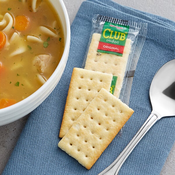A bowl of soup with a spoon and Kellogg's Club Crackers on a blue cloth.