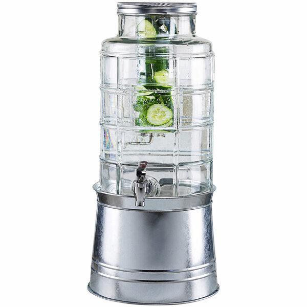 A Stylesetter glass beverage dispenser with a galvanized metal base filled with water and cucumbers.