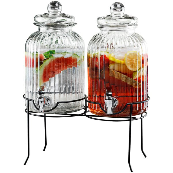A Stylesetter glass beverage dispenser with black metal stand holding water and fruit.