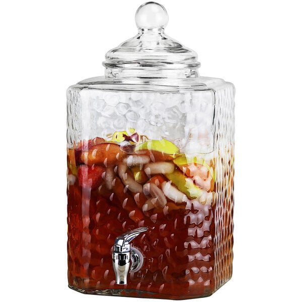 A Stylesetter hammered glass beverage dispenser filled with a drink and fruit on a counter.