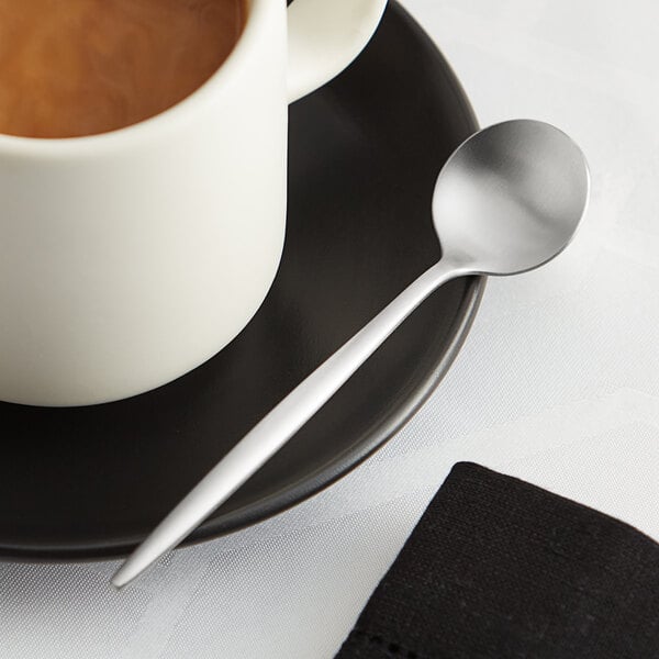 An Acopa brushed stainless steel demitasse spoon on a saucer with a cup of coffee.
