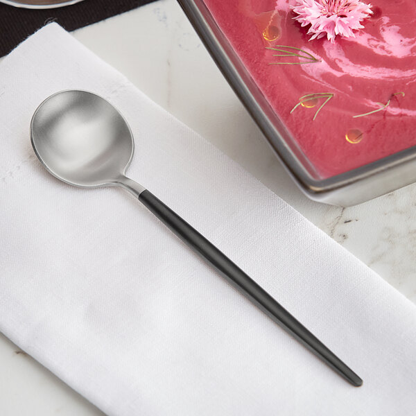 An Acopa brushed stainless steel bouillon spoon on a napkin next to a bowl of red soup.