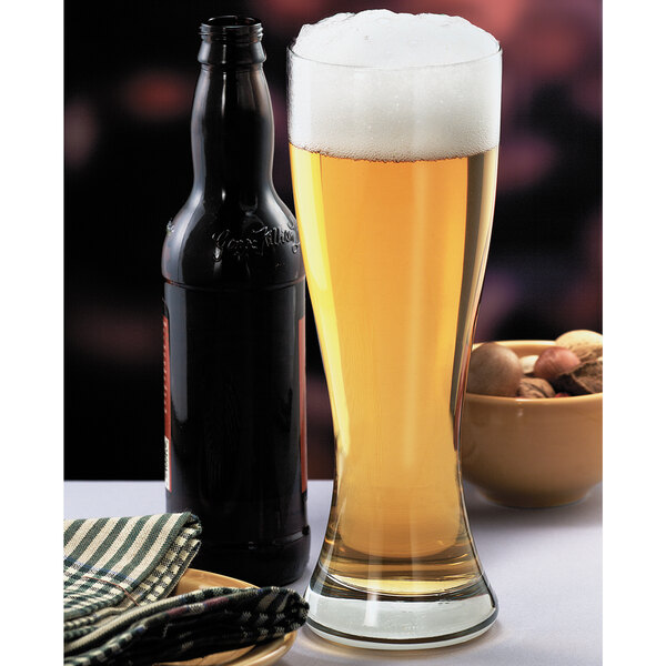 A Libbey giant pilsner glass filled with beer next to a bottle of beer.