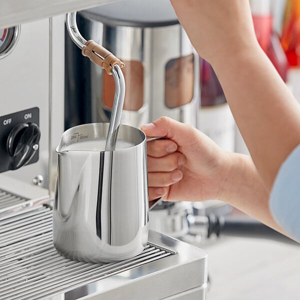 A person using an Estella Caffe stainless steel frothing pitcher to pour milk into a cup.