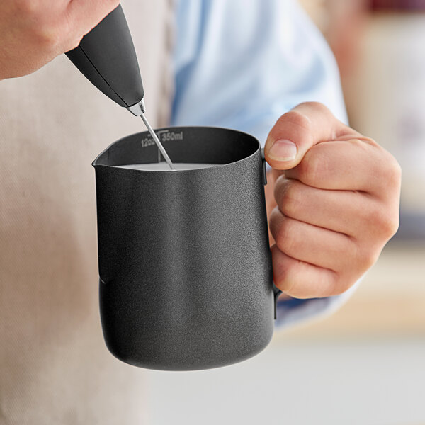 A hand using an Acopa black frothing pitcher to pour milk into a mug.