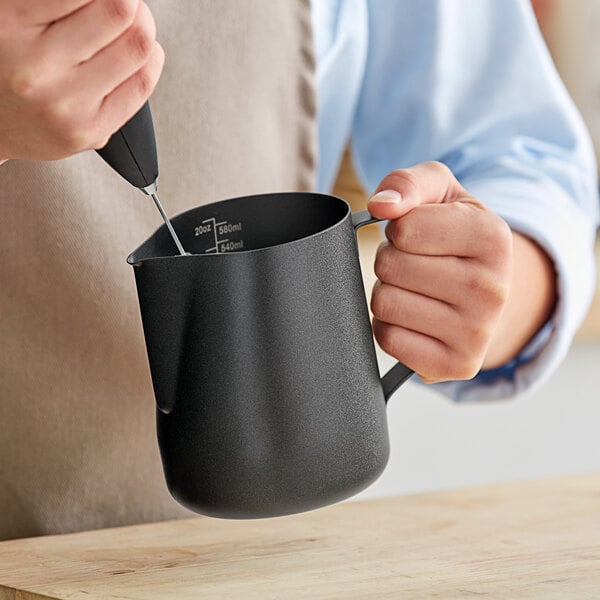 A person using an Acopa black frothing pitcher to pour milk.