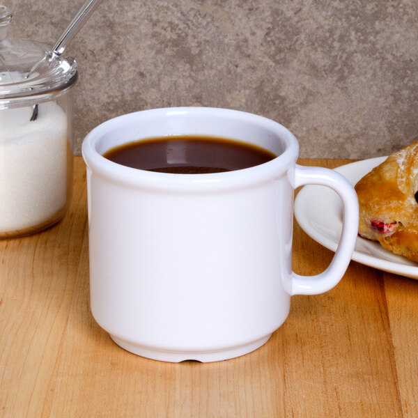 A white plastic mug filled with brown liquid on a table with a pastry.