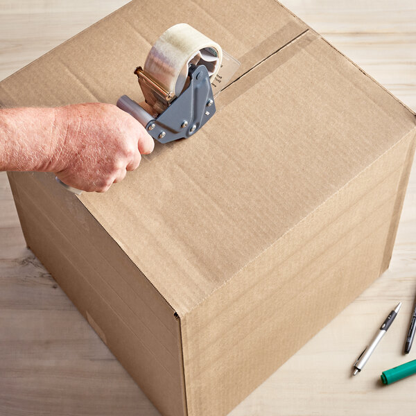 A hand using a tape dispenser to open a Lavex Kraft shipping box.