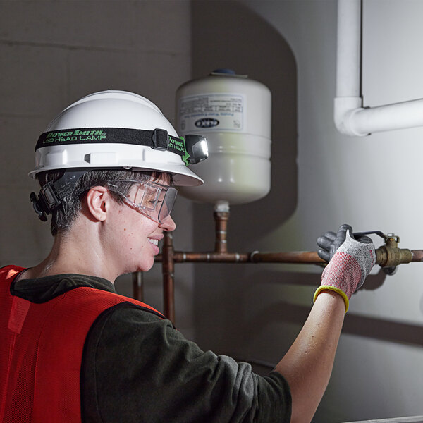 A woman wearing a hard hat and goggles using a PowerSmith LED headlamp to work on a pipe.