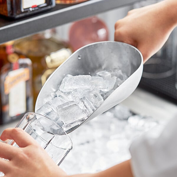 A hand using a Choice aluminum scoop to pour ice into a glass on a counter.