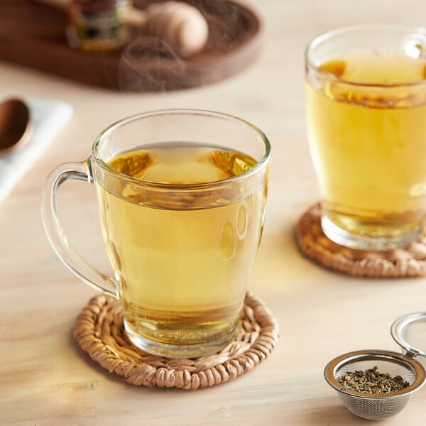 A glass mug of Davidson's Organic Peppermint Leaves tea on a wooden table with a strainer of herbs.