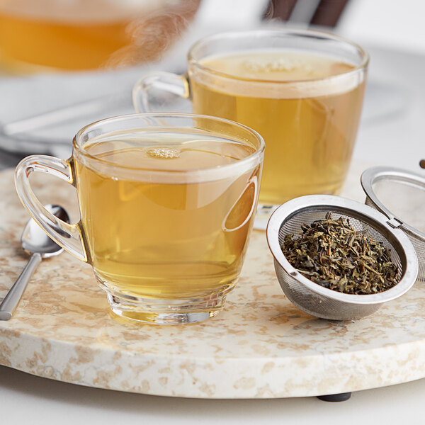 A glass cup of Davidson's Organic Moroccan Green with Mint tea next to a strainer filled with green leaves.