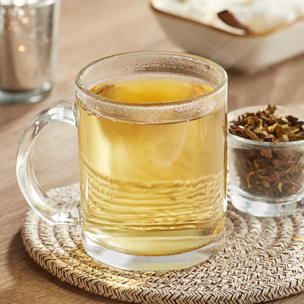 A glass mug of Davidson's Organic White Spicy Peach Tea on a table next to a bowl of dried herbs.
