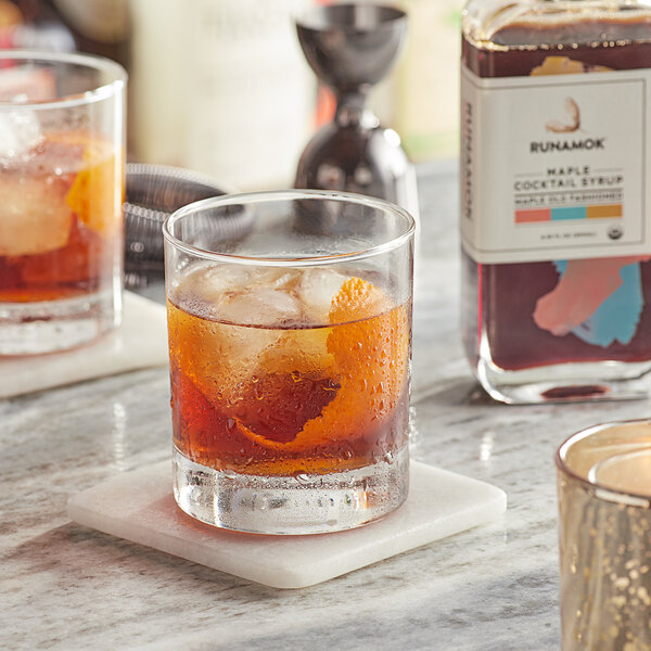 A glass of Runamok Maple Old Fashioned Cocktail with ice and orange slices on a marble table.