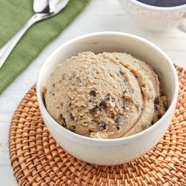 A bowl of The Cookie Dough Cafe Oatmeal Chocolate Chip Cookie Dough with a spoon next to a bowl of ice cream and a cup of coffee.