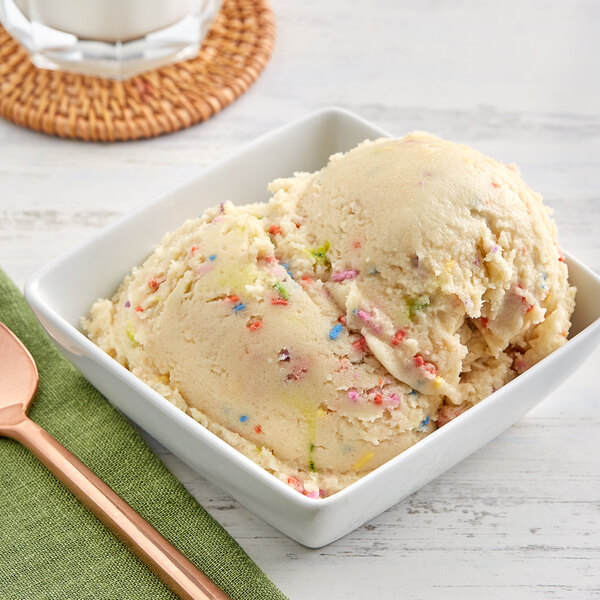 A bowl of ice cream with The Cookie Dough Cafe Edible Confetti Cake Cookie Dough on top and sprinkles with a spoon.