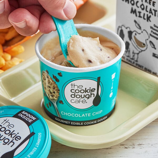 A hand holding a spoon over a mini cup of The Cookie Dough Cafe chocolate chip cookie dough.