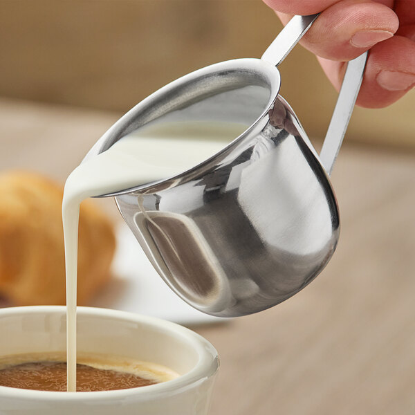 A person pouring milk into a cup of coffee using a stainless steel bell creamer.