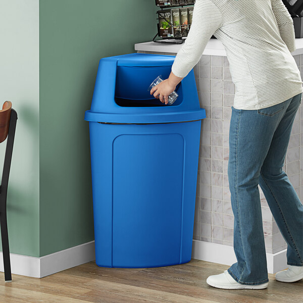 A woman using a Lavex blue corner round trash can with a blue push door lid to throw away a plastic cup.