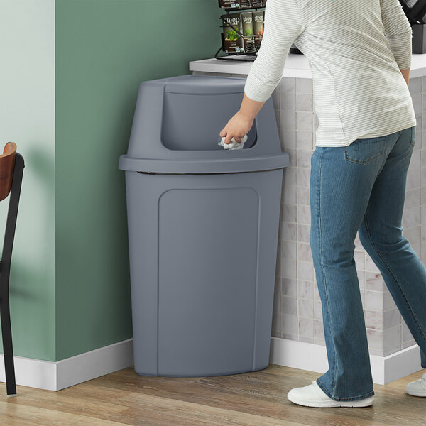 A woman in a white shirt and jeans using a Lavex Gray Corner Round trash can with a gray push door lid.