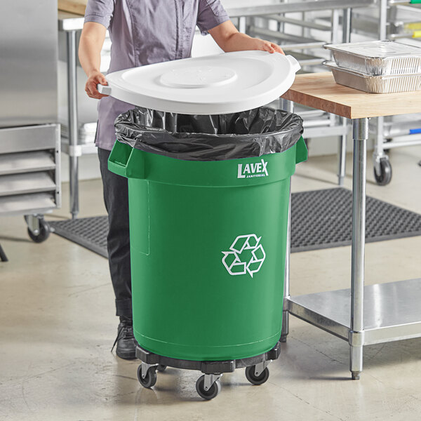 A woman in a school kitchen putting a white lid on a green Lavex recycling can.