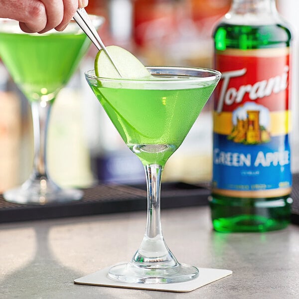 A person pouring Torani Green Apple flavoring syrup into a glass of green liquid.