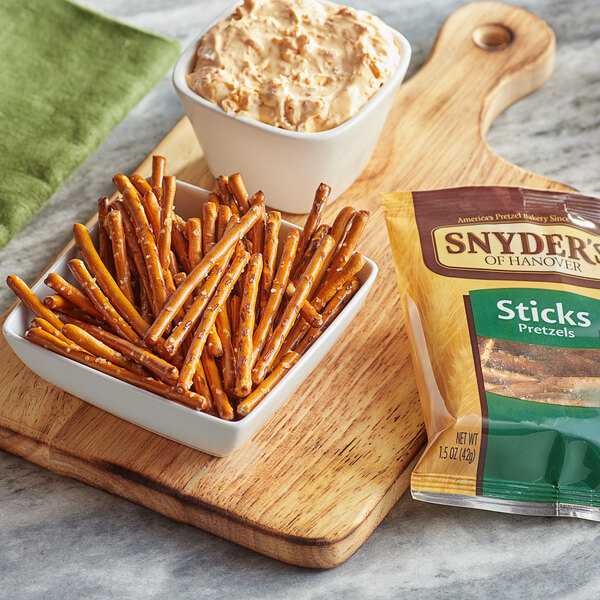 A bowl of Snyder's of Hanover Pretzel Sticks and dip on a wooden cutting board.