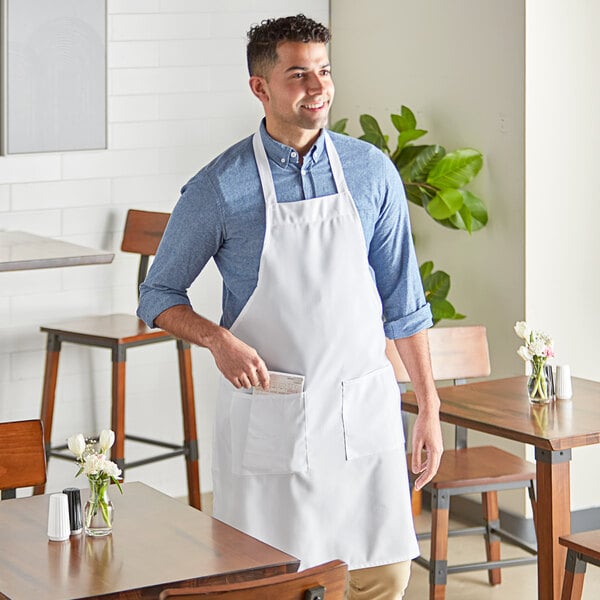 A man wearing a white Choice standard bib apron with 2 pockets standing in a professional kitchen.