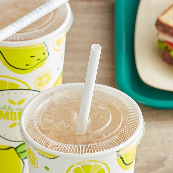 A plastic cup with a SOFi white paper straw in it next to a sandwich on a table.