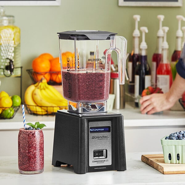 A Blendtec Connoisseur commercial blender with a jar of red liquid and a blueberry smoothie in front of a person.