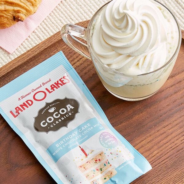 A glass of Land O Lakes Cocoa Classics with whipped cream next to a packet of food.