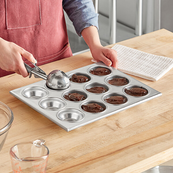 A woman using a spoon to put brown cupcake batter into a Choice aluminum muffin pan.