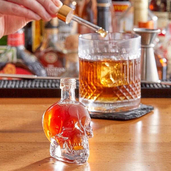 A person pouring liquid into an Acopa glass skull bitters bottle on a counter in a cocktail bar.
