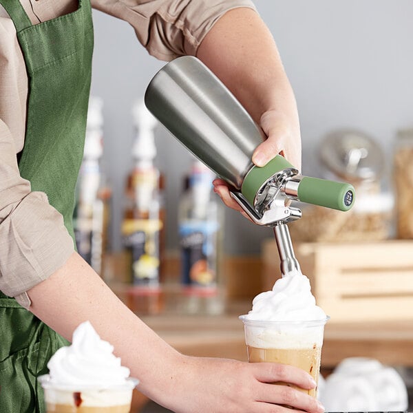 A person using a green stainless steel iSi whipped cream dispenser to pour whipped cream on a drink.