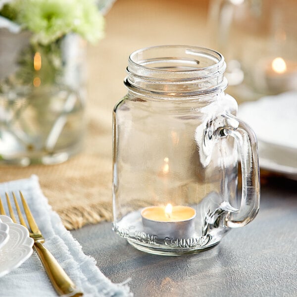 An Acopa mason jar candle with a handle on a table with a lit candle and a gold fork.