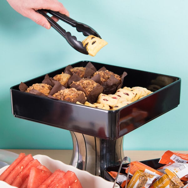 A person using tongs to serve food from a black square deli crock.