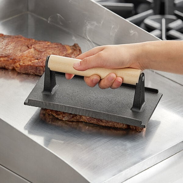 A hand using a wooden-handled cast iron steak weight on a grill.