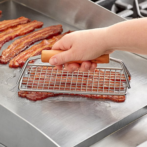 A hand using a Choice wood handle bacon press over a rack of bacon.