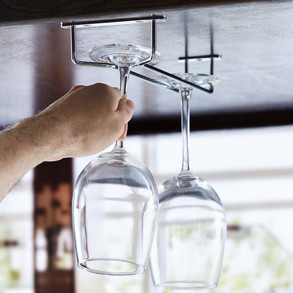 A person using a Regency Chrome Plated Glass Hanger Rack to hang wine glasses over a counter.