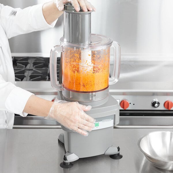 A woman in gloves using a Waring food processor with a clear bowl on a counter in a professional kitchen.
