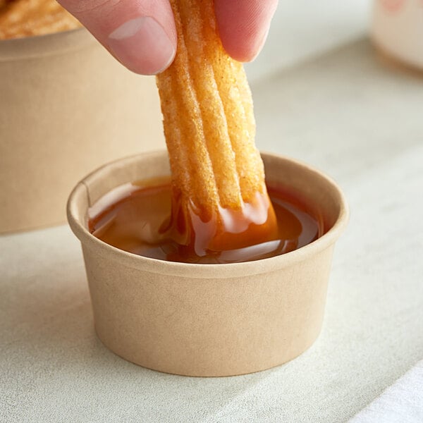 A hand dipping a Choice kraft paper take-out container of churros into a cup of liquid.