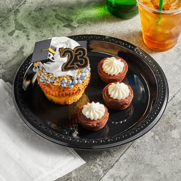 A Huhtamaki Chinet black plastic plate with a cupcake with white frosting and a drink on a table with cupcakes.