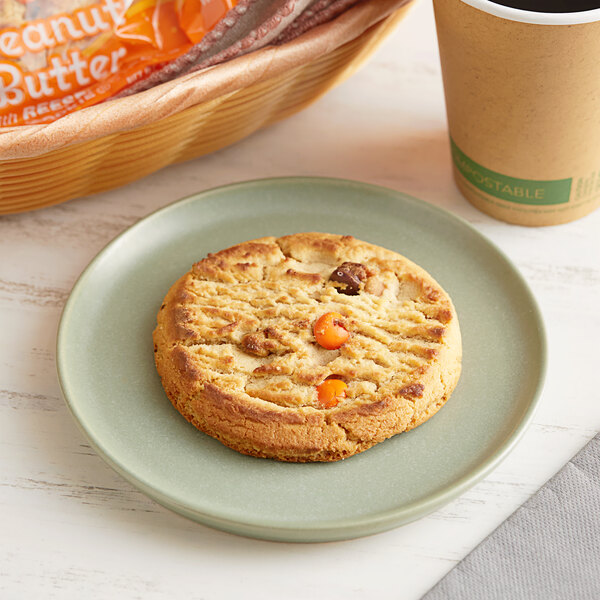 An individually wrapped Best Maid peanut butter cookie with Reese's Pieces on a plate next to a cup of coffee.